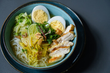 A bowl of noodles with eggs, slice of chicken and vegetable as  a background