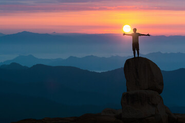 Hiker with backpack seeing the sunrise from a mountaintop while holding up his hands.