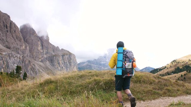 Arrival. Young man hiker hiking on mountain dirt road with cloudy sky and fog. Yellow jacket, backpack, black beard and beanie. Traveling Dolomites, Italy.