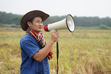 Asian man farmer wears hat, holds megaphone at agriculture field. Concept, communication , announce. Protest. Protester. Bullhorn public address. Loudspeaker technology for outdoor announcement.