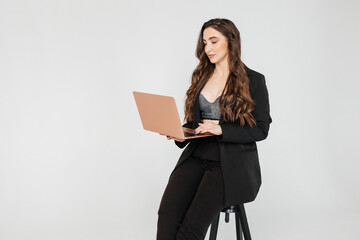 young girl with long dark curly hair in a black suit with a laptop sits on a white background. Businesswoman working online