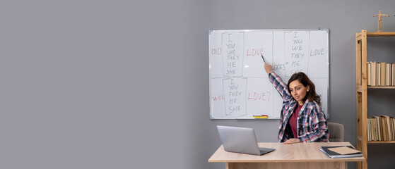 Female English teacher sitting at table, pointing at whiteboard, explaining grammar rules, having video conference with students. Education and e-learning concept.