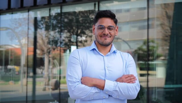 close up portrait of folded hispanic young entrepreneur smiling, looking at camera at workspace. Front view of one latin happy employee office crossed arms worker with glasses and successful attitude
