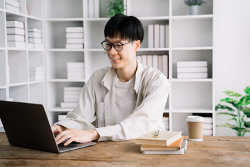 male student in casual clothes sitting at wooden table with laptop and writing notes while preparing for exam in library.