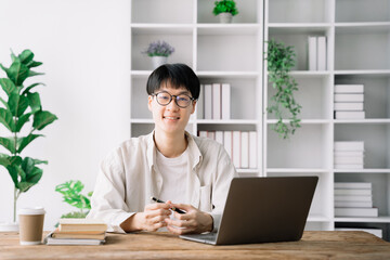 male student in casual clothes sitting at wooden table with laptop and writing notes while preparing for exam in library.