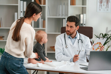 Fototapeta premium Having consultation. With baby and mother. Doctor in formal clothes is working in the cabinet
