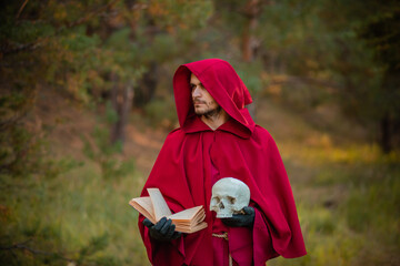 A sorcerer in red clothes with a skull and a book in his hands reads a spell. Medieval monk reads a prayer. - obrazy, fototapety, plakaty