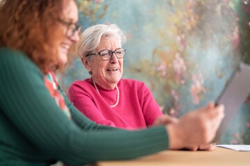 Cheerful gray haired woman in glasses and redhead daughter sitting at table while using tablet making a video call