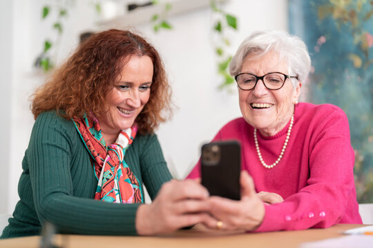 Smiling mature redhead female sitting at table with gray haired mother in pink sweater using mobile phone watching funny photos