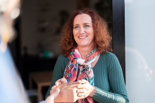 Positive mature redhead haired woman in glasses and pink sweater receiving package from courier while standing at doorway