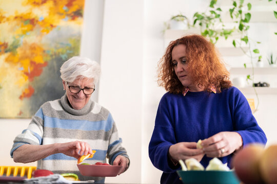 Mature curly haired woman and aged mother preparing healthy food in kitchen while adding broccoli to salad and watching tutorial on laptop