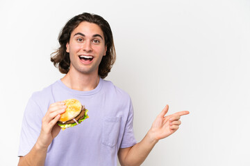 Young handsome man holding a burger isolated on white background surprised and pointing finger to the side