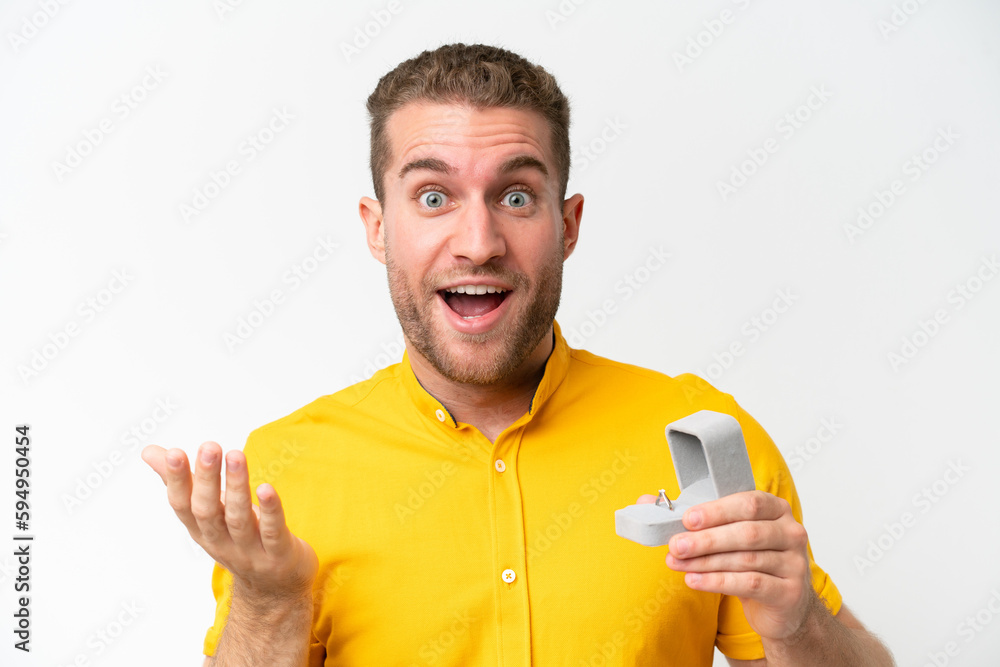 Wall mural Young man holding a engagement ring isolated on white background with shocked facial expression