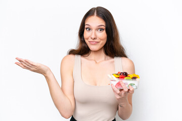 Young caucasian woman holding a bowl of fruit isolated on white background having doubts while raising hands