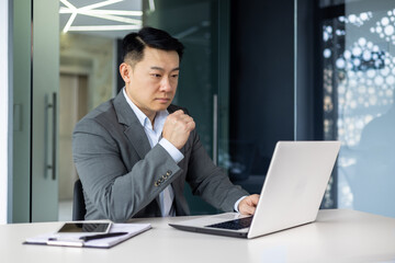 Fototapeta premium Serious thinking asian businessman working inside office sitting at table with laptop, boss making important financial decision, asian mature man in business suit at workplace.