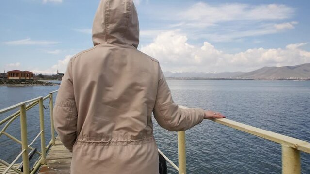 a woman in a sand-colored jacket, wearing a hood, walks along the pier on the shore of a mountain lake, holding on to the handrails of the pier. Young adult woman enjoying sea and mountains landscape