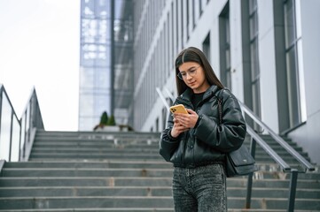 Standing on the stairs. Young woman is outdoors near building is holding smartphone