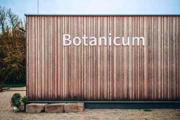 Wooden facade of small rectangular building with lettering Botanicum on the wall outdoors in the garden and blue sky line on the background.