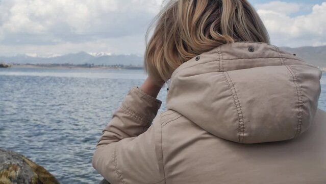 A young blonde adult woman sits on the shore of a cold mountain lake and looks thoughtfully into the distance at high mountains with snowy peaks. Enjoying nature and relaxing alone