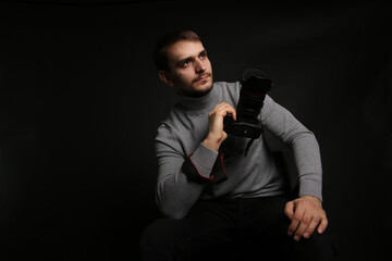 Portrait of a young handsome man in a gray jacket with a camera in his hand. The photographer on a black background sits on a chair.