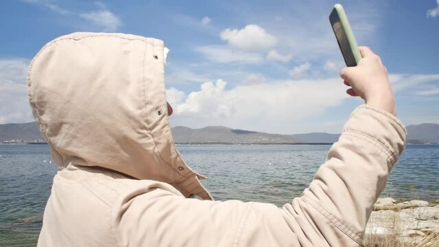 An adult young woman in a jacket and hood takes a selfie holding a smartphone in front of her at arm's length standing on the shore of a mountain lake and photographing herself with the landscape