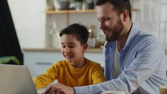 Father helps son with homework. Dad helping kid with online classes at home. Man and boy use laptop