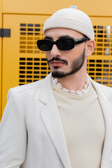 Portrait of fashionable gay man in hat and sunglasses standing on urban street.