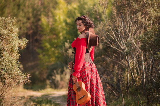 A young woman with dark country style hair in a red peasant jacket and plaid skirt holding a mini guitar.Playing the ukulele instrument in nature