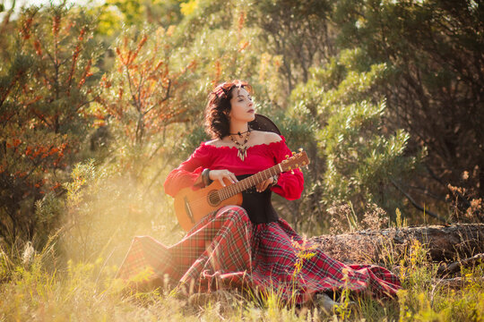 A young woman with dark country style hair in a red peasant jacket and plaid skirt holding a mini guitar.Playing the ukulele instrument in nature