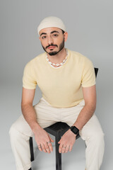Fashionable gay man in hat looking at camera while sitting on chair on grey background.