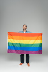 Full length of young gay man holding lgbt flag and smiling at camera on grey background,...