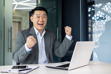Portrait of successful asian boss, businessman celebrating results achievement, man in business suit smiling and looking at camera holding hands up triumph gesture, mature investor inside office.