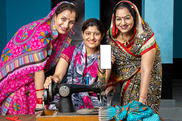 Portrait of three happy traditional indian women wearing sari using sewing machine while showing smart phone with empty display to put advertisement, blank android phone screen.