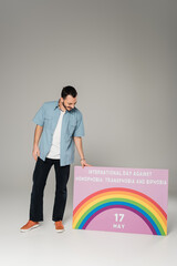 Full length of smiling gay man standing near placard with International Day Against Homophobia, Transphobia and Biphobia lettering on grey.