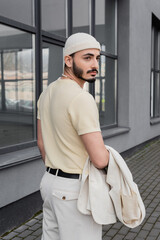 Fashionable gay man in hat holding jacket near building outdoors.