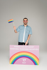 Cheerful gay man holding lgbt flag near placard with International Day Against Homophobia,...
