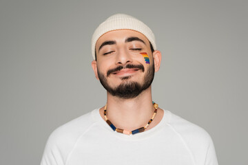 portrait of smiling gay man in hat with lgbt flag on cheek standing isolated on grey.