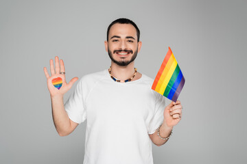 Smiling gay man with lgbt flag and heart on hand looking at camera isolated on grey, International...