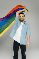 Young homosexual man in medical mask holding lgbt flag isolated on grey, international homophobia...