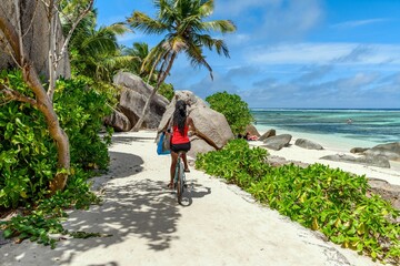 Woman cyclist riding a bicycle along a sandy pathway in tropical beach on La Digue island