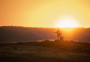 Silhouette of a man playing guitar at sunset in the field. Hills in the background. Orange colored sky.
