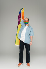 Full length of young gay man in medical mask holding lgbt flag on grey background.