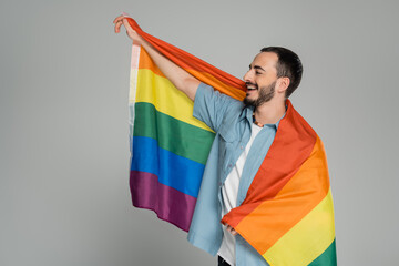 Bearded and cheerful gay man holding lgbt flag and looking away isolated on grey, International Day...