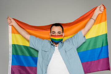 Young gay man in medical mask holding lgbt flag isolated on grey, International Day Against...