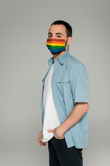 Brunette gay man in medical mask with lgbt flag looking at camera isolated on grey.