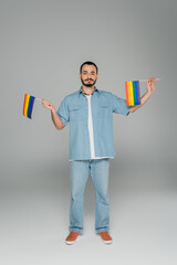 Full length of young gay man holding lgbt flags while standing on grey background, International...