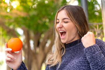 Young pretty Romanian woman holding an orange at outdoors celebrating a victory