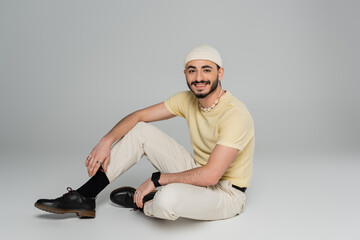 Cheerful gay man in hat looking at camera while sitting on grey background.