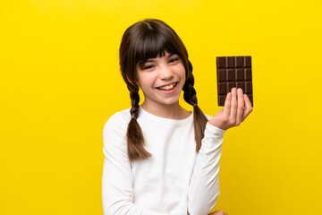 Little caucasian girl isolated on yellow background taking a chocolate tablet and happy