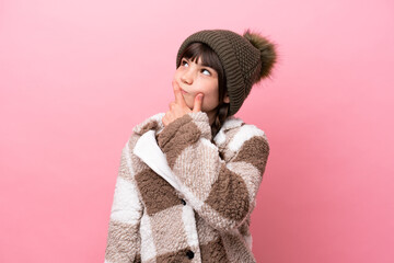Little caucasian girl with winter jacket isolated on pink background having doubts and with confuse face expression
